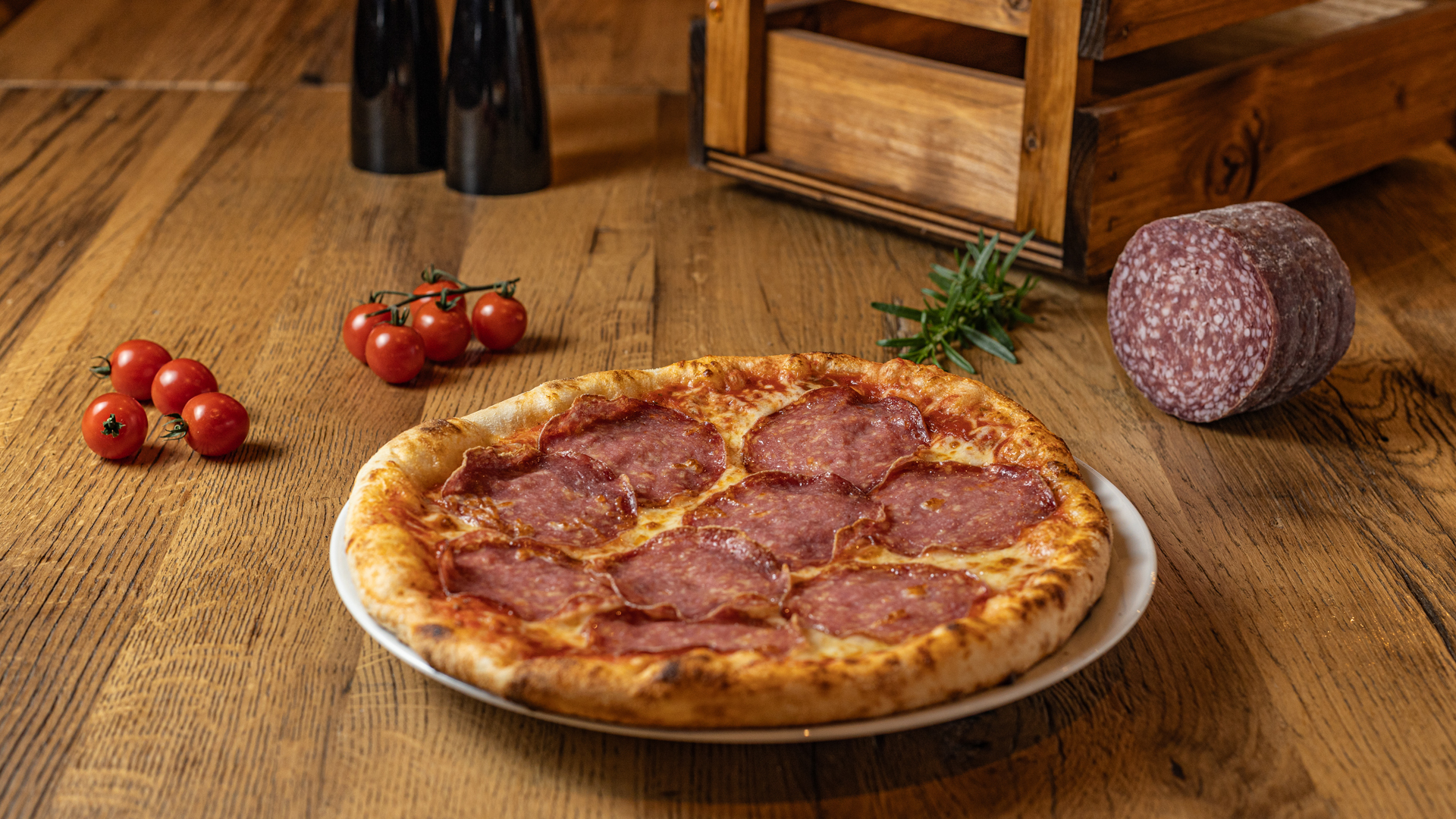 A pepperoni pizza sits on a white plate on a wooden table, surrounded by cherry tomatoes, a sprig of rosemary, a whole salami, and two black bottles.
