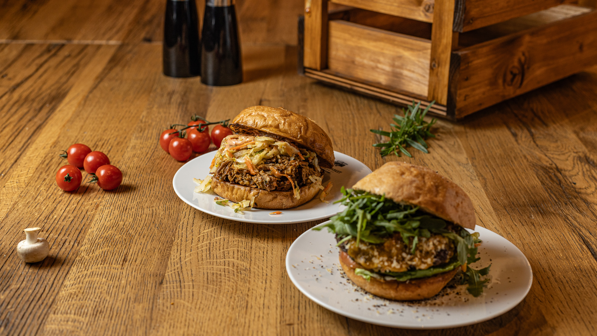 Two gourmet burgers on white plates sit on a wooden table, one topped with shredded meat and cheese, the other with arugula and seeds. Cherry tomatoes and a small mushroom are nearby, with salt and pepper shakers in the background.