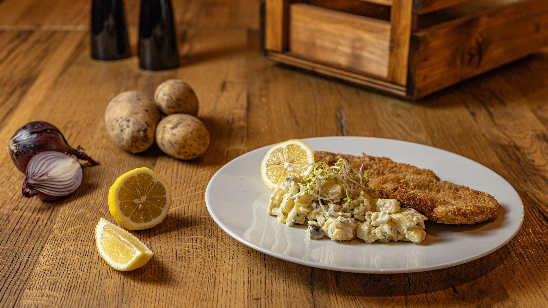 A plate with breaded schnitzel, potato salad, and lemon slices on a wooden table, surrounded by raw potatoes, a sliced red onion, lemon wedges, salt and pepper shakers, and a wooden box in the background.