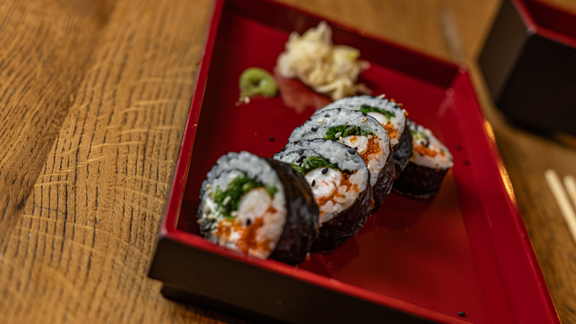 Six sushi rolls topped with orange roe are neatly arranged in a red bento box on a wooden table, accompanied by wasabi and pickled ginger in the corner.